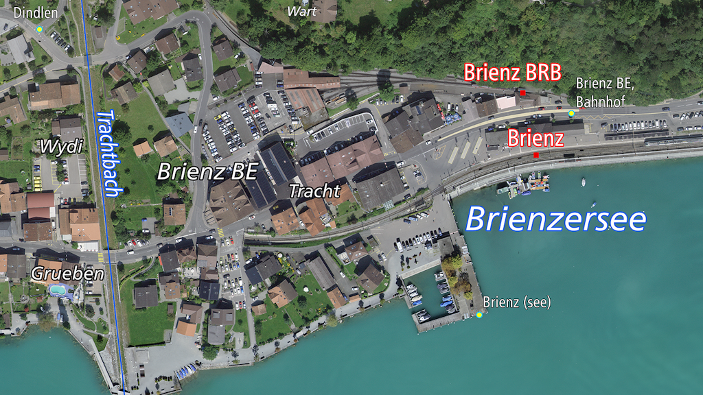 L'immagine mostra una veduta aerea di Brienz BE con il paese, la stazione ferroviaria e il lago di Brienz. 