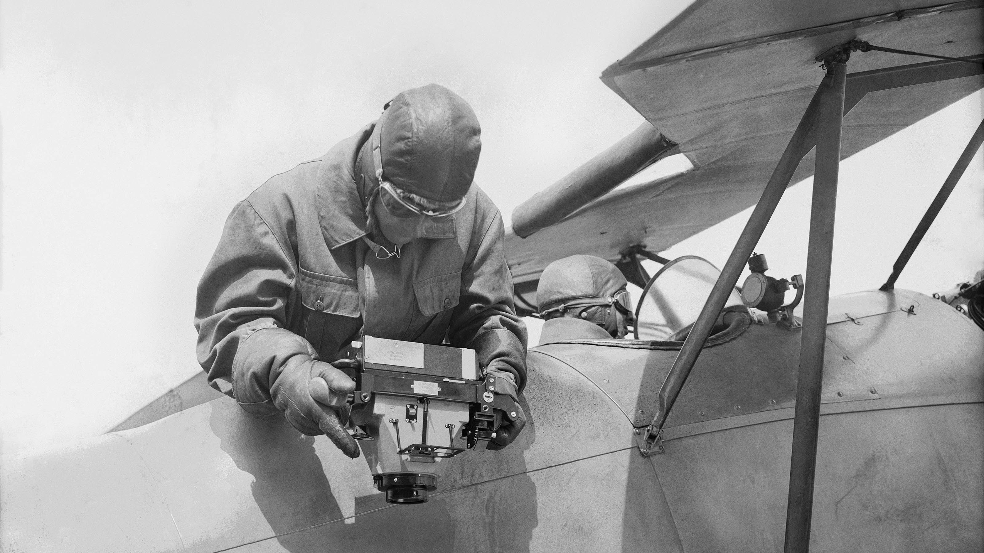 A pilot sits at the controls of an aircraft while the observer holds the aerial camera by hand overboard and photographs downwards.