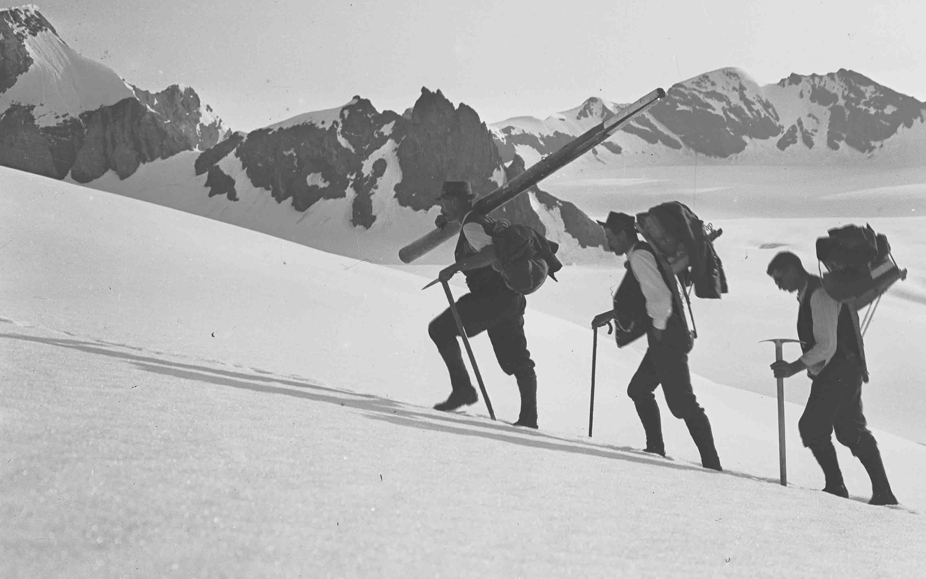 Trois géomètres en train de gravir une montagne sur un champ de neige. Les trois hommes sont lourdement chargés de bagages.