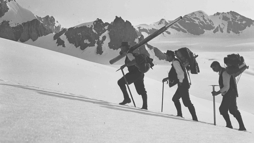 Trois géomètres en train de gravir une montagne sur un champ de neige. Les trois hommes sont lourdement chargés de bagages.
