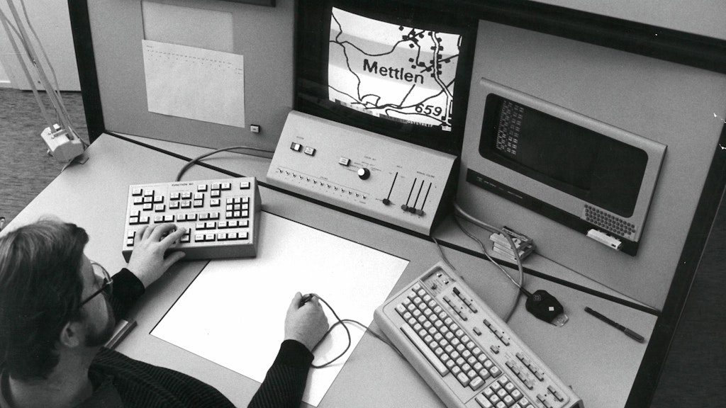 A swisstopo cartographer operating the office's first digital mapping system, SciTex. The cartographer works on two screens and several keyboards and control panels. In the center of the picture is a touch panel and the cartographer's electronic pen, which he can use to transfer information to the computer.