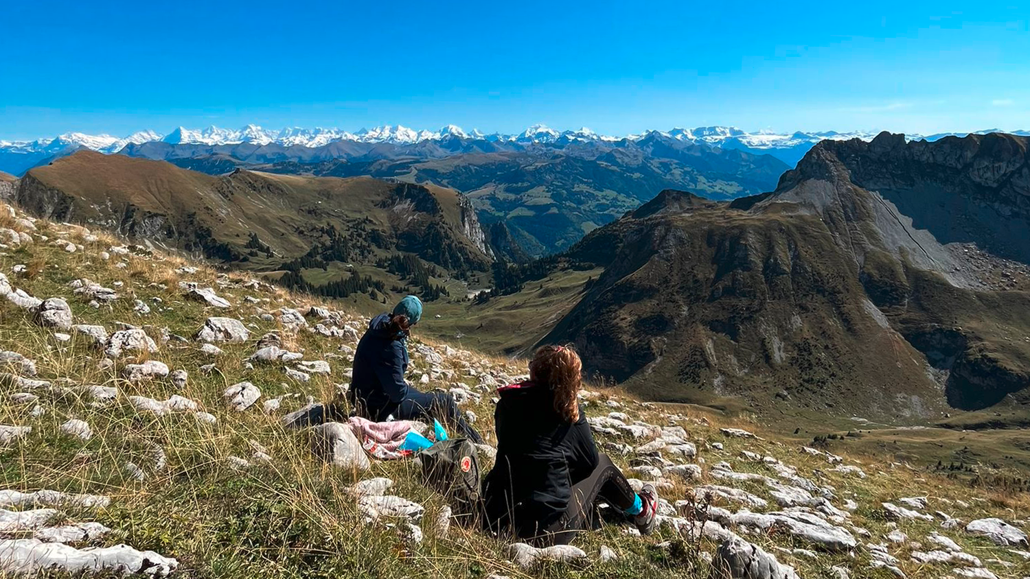 Bei schönstem Wetter machen zwei Wanderinnen Rast auf der Kaiseregg und blicken über die Berglandschaft in Richtung Alpen. Im Hintergrund wolkenloser, blauer Himmel.