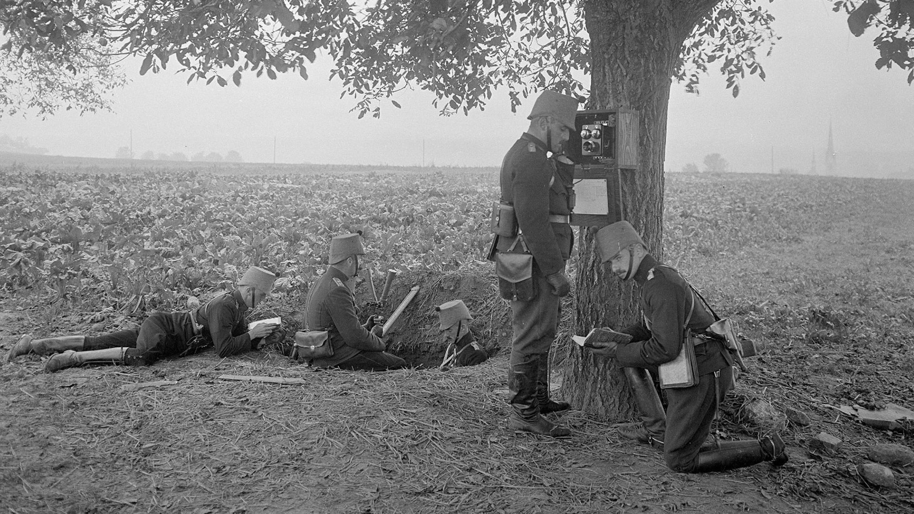 Vier Soldaten bei einem Acker. Die Soldaten sind teilweise in einem Schützengraben. Neben ihnen steht ein Kommandant, der in ein Telefon spricht, das an einem Baum angebrachten ist.