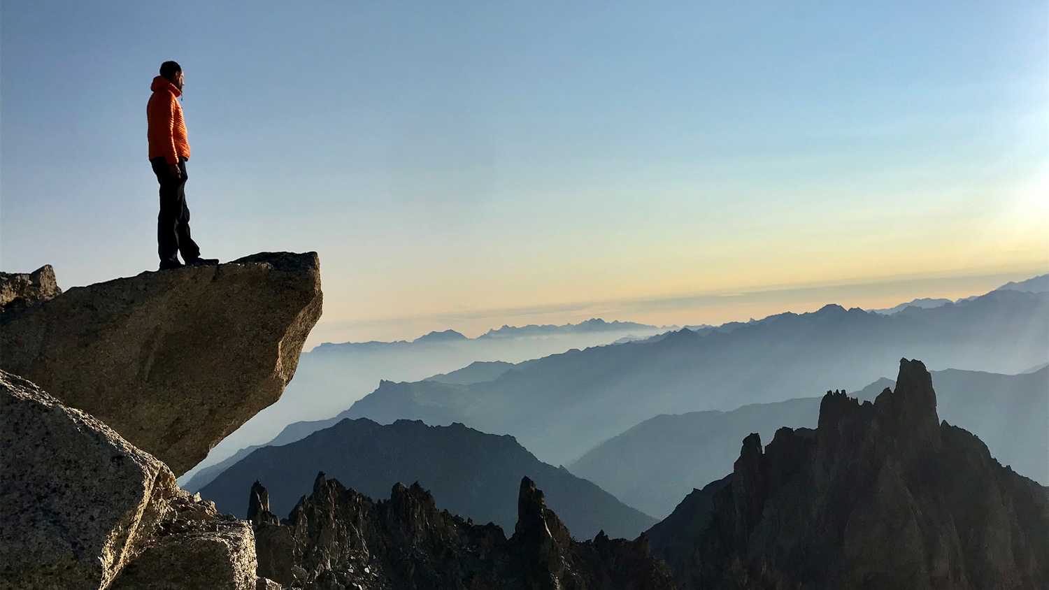 Hügelige Landschaft im Vordergrund steht Wanderer auf Steinvorsprung und blickt in die Ferne über die Berglandschaft