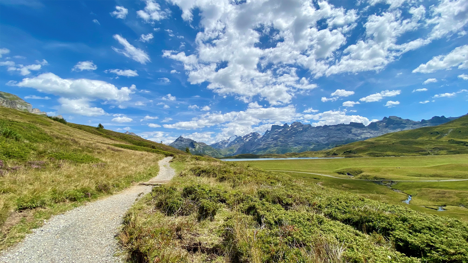 Grüne Landschaft in Melchsee-Frut. links im Bild schöner Weg mit Naturbelag, im Hintergrund Berge, blauer Himmel mit einigen zerstreuten Wolken