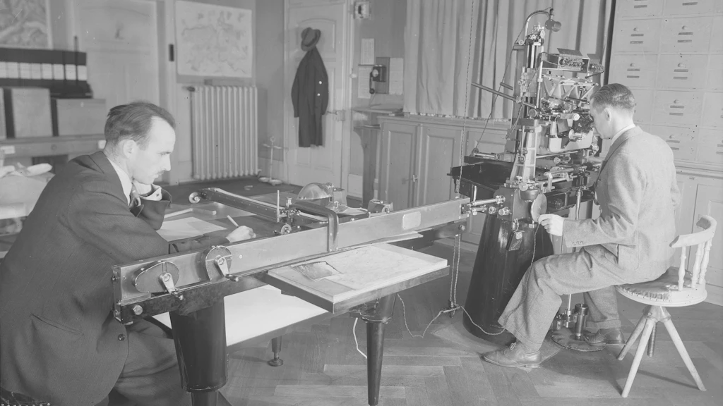 Travail sur le stéréoautographe Wild n° 3, 1938 : l’opérateur regarde une paire de photographies à travers un stéréoscope. À l’aide de deux manivelles et d’une pédale, il actionne l’appareil.