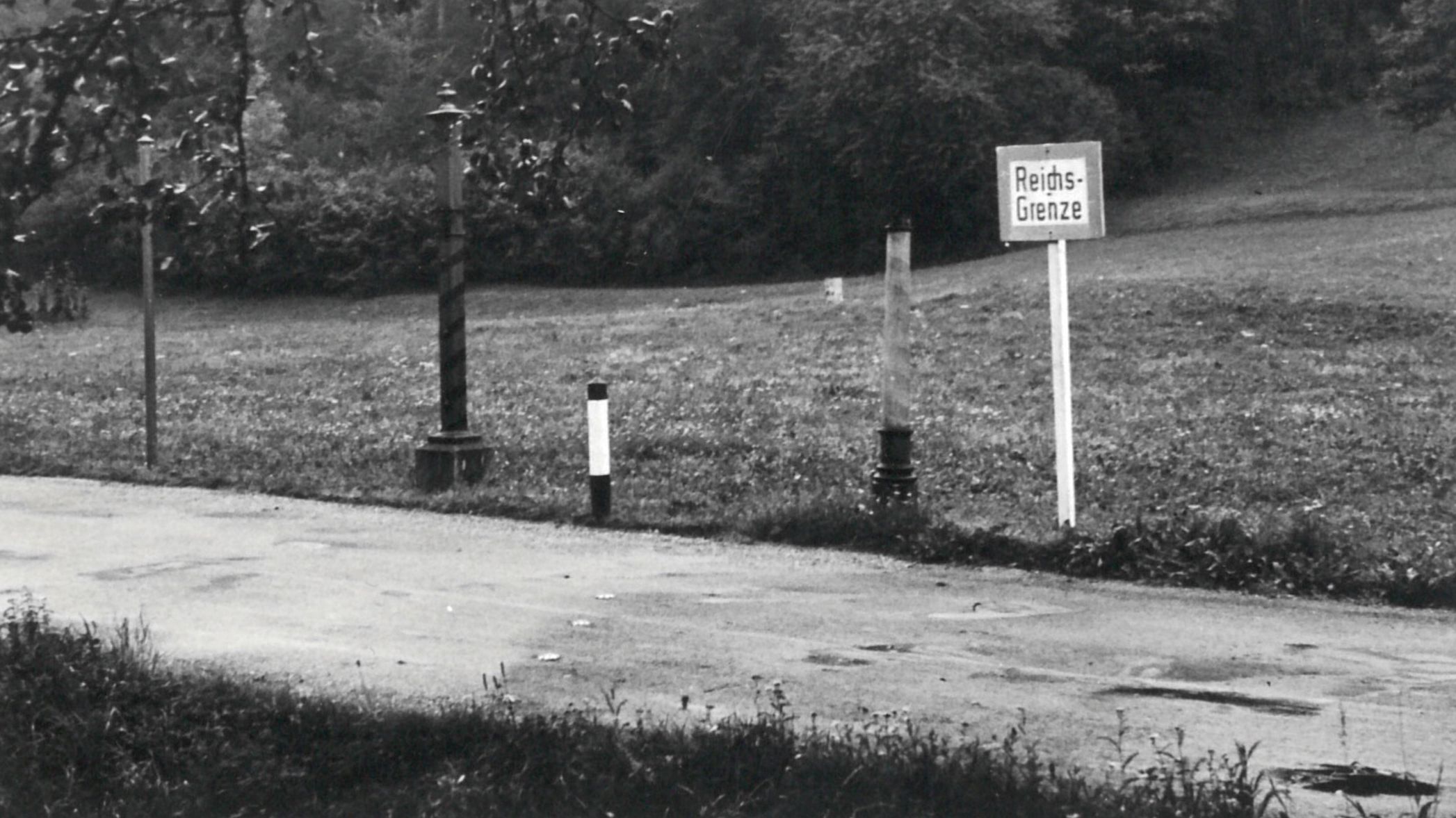 Photo en noir et blanc avec rue, borne et panneau de bordure : frontière du Reich